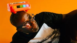 Charles Joseph Smith tilts his head toward the camera, balancing a colorful toy instrument on top of his head, wearing dark-framed glasses and a black jacket. Photo by Dennis Larance.