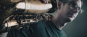 Ethan Helm holds a saxophone resting on his shoulder, looking slightly off-camera. Close crop, muted teal background. Tortoiseshell glasses.