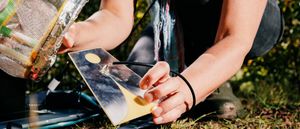 Lisa Ann Schonberg crouches outdoors on grass, handling a small reflective metal plate with a circular cutout, cables, and a foil-wrapped bundle of sound equipment.