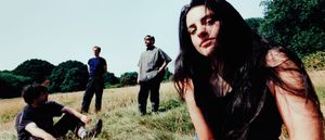 One member of Modern Woman leans close to the camera, long dark hair windswept across her face, while three others stand and sit in a sun-bleached field behind them.