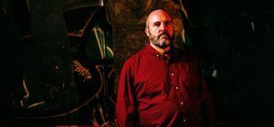 Nick Fraser stands against a dark wall covered in graffiti, wearing a deep red button-down shirt, arms at his sides, with a full beard and a steady, neutral gaze toward the camera.