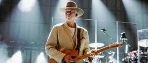 Phil Manzanera performs onstage holding a Fender Telecaster, wearing a cream suit and wide-brimmed hat, lit by dramatic stage lighting against an arched industrial backdrop.