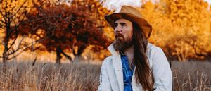 Bob Wagner stands in a dry autumn field, long hair and beard, wearing a tan wide-brim hat and white jacket over a denim shirt, gazing to one side against a backdrop of amber-lit trees.