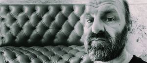 Chik White, bearded, gazes into the camera in a black-and-white close-up, seated against a tufted leather couch with a cracked plaster wall behind him.