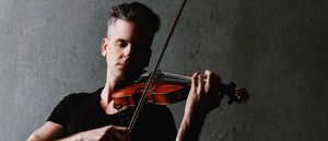 Christopher Tignor plays violin, eyes downcast, against a textured gray wall. He wears a black t-shirt, with a tattoo visible on his left arm.