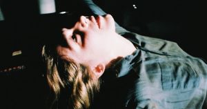 Eliana Glass reclines with head tilted back and eyes closed, shot on grainy film against a dark background, a piano visible at the left edge of the frame.