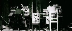 Black-and-white photo: two performers sit with backs to camera, each facing a rack of vintage electronic equipment. A large unit with a VU meter stands between them on a bare stage.