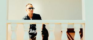 Mark Barrott stands behind a white balustrade, wearing dark sunglasses and a black jacket, gazing to one side against a pale, sun-bleached wall.