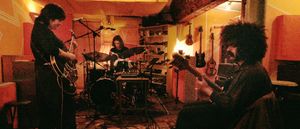 Myra Lee rehearse in a warmly lit studio, guitarist and bassist flanking a drummer at center, guitars hanging on the wall behind them.