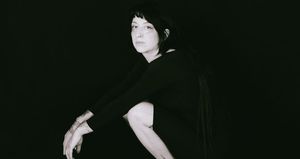 Ora Cogan stands against a black background in a black t-shirt, glancing over one shoulder, tattooed forearms crossed in front of her. Black and white.