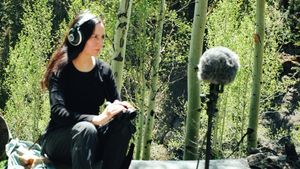 Patricia Wolf, dressed in black, crouches with headphones on, listening intently, while a boom microphone on a tripod faces them amid sunlit aspen trees.