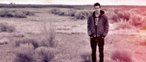 Phil Geraldi stands alone in an open desert scrubland, hands in pockets, wearing a hoodie and dark jeans. The photo has a faded quality with warm red-pink tones bleeding in from the right.