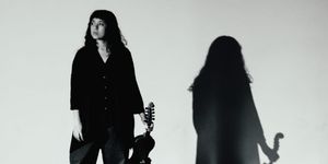 Zosha Warpeha stands holding a violin at her side in a stark black-and-white studio shot, her dramatic shadow cast large on the wall behind her.