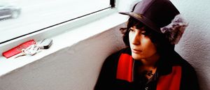 zzzahara seated against a wall beneath a window, wearing a fur-eared cap and red-and-black jacket, gazing upward. Keys and earbuds rest on the sill behind them. Photo by Lindsey Byrnes.
