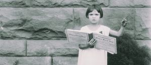Lorraine Miles holds open copies of the "Original Sacred Harp" hymnal before a stone wall, one arm raised, in a vintage black-and-white photograph.