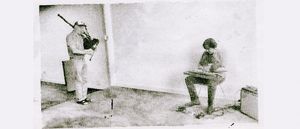 David Watson stands playing Highland bagpipes near a corner, while Bill Nace sits across the room playing a taishōgoto on his lap, in a grainy halftone photograph.