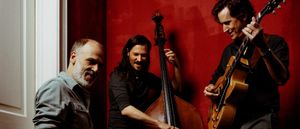 Three members of Cento Threeo against a deep red wall: one leans against a doorframe at left, one plays upright bass at center, one plays archtop guitar at right.