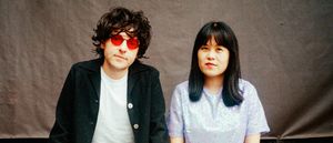Bryce Barsten and Anne Tong of Chinese American Bear seated against a grey backdrop, Barsten in red tinted round sunglasses and a black jacket, Tong in a lavender sequined top.