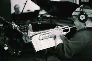 Dave Douglas plays trumpet in a recording studio, reading from sheet music on a stand, headphones on, a vintage microphone inches from the bell.