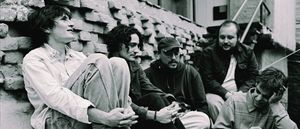 Diles que no me maten sit together against a brick wall in black and white, five members leaning and talking, one resting his head on his hand.
