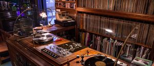 Inside jazz kissa Disk: two turntables and a vintage mixer on a wooden counter, floor-to-ceiling shelves of vinyl covering the walls behind, a brass lamp casting light over the setup.