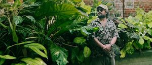M. Geddes Gengras stands among towering tropical plants with broad green leaves, wearing a cap, round sunglasses, and a patterned shirt, a brick wall and white birdcages behind him.