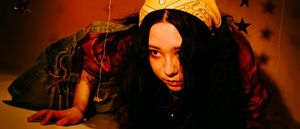 Mixol crawls toward the camera along the floor, hair falling over one eye, in a denim jacket and yellow patterned headscarf, against an orange wall hung with dark stars.