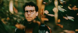 Scrimshire photographed among autumn woodland foliage, wearing glasses and a patterned knit sweater, looking directly into the camera.