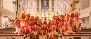 Dozens of The Goddess Party performers in orange wigs and pink and gold sequins pose across the pews of an ornate church, arms raised, 'GODDESS PARTY' projected on the wall behind them.