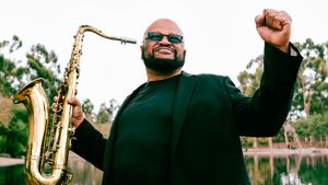 Azar Lawrence stands beside a lake holding a tenor saxophone in one hand, the other fist raised, in a black jacket and sunglasses.
