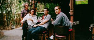 Bondo sit at an outdoor bamboo bar ringed with tropical plants, all four turning to face the camera, a soccer match playing on a screen above.
