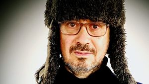 David Norland in a thick fur trapper hat with ear flaps and clear-framed glasses, looking directly at the camera against a pale grey wall.