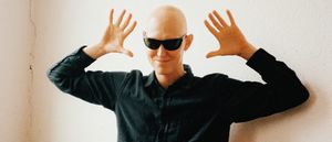 GB stands against a white wall in a black shirt and dark sunglasses, both hands raised palms-out beside his head.