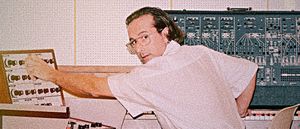 Jolly Mare, in glasses, reaches to turn a knob on a mixing panel, a large modular synthesizer mounted on the wall behind him.