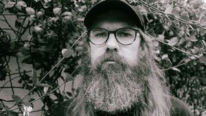 Josh Mason looks straight at the camera from behind leafy vines, face filling the frame, in a dark cap and round glasses, his long beard reaching the bottom edge. Black and white.