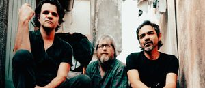 Riviera Gaz seated in a narrow alley with peeling walls — Gustavo Riviera at left with a raised fist, Steve Shelley centre in a plaid shirt, Paulo Kishimoto at right in a black T-shirt.