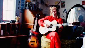 Sophie Wellington sits playing a pale acoustic guitar and laughing, dressed in red, in a warm cluttered room with an archtop guitar leaning beside her and light from a window.