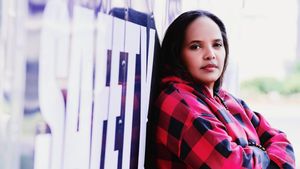 Terri Lyne Carrington leans against a window in a red and black plaid hoodie, arms folded, looking directly at the camera past the word SAFETY in large white letters on the glass.