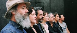 The Odyssey Cult in a row against a black wooden wall, all in profile facing right, Ethan Miller nearest the camera in a pale hat and denim jacket, long grey beard.