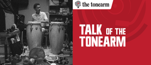 Irán Farías Saínz sits behind two congas in a cluttered studio ringed with tambourines, shakers, and cowbells. A red panel beside him reads TALK OF THE TONEARM.