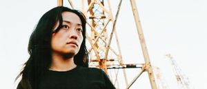 sachi's mirror stands below a rusted industrial crane, shot from a low angle against a pale sky, dark hair blown across one shoulder, looking off past the camera.