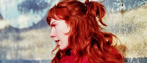 Elanor Moss in profile, red hair pulled into a high ponytail with a fringe, wearing a red top, lips parted. Behind her a blurred backdrop of pale blue and weathered cream.