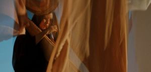 Lara Somogyi plays a harp behind drifting sheets of amber fabric, her face and hands glimpsed through the strings against a pale blue backdrop.