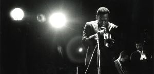 Miles Davis bends toward the mic under two bright stage lights, Wynton Kelly at the piano and Paul Chambers on bass behind him in the dark.