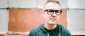 Jeff McIlwain, aka Lusine, in a teal hooded jacket and black glasses, facing the camera in front of a wall of terracotta panels edged in pale grey.