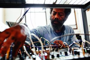 Robert Aiki Aubrey Lowe looks down at a modular synthesizer, framed by its patch cables and knobs in the foreground, window light behind him, a blurred hand reaching in from the left.