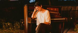 Sova sits beside an old wooden upright piano in a backyard at dusk, its hammers exposed, head resting on one hand and smiling downward, lit warm against the dark garden.