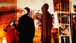 Jon Hassell, seen from behind with trumpet in hand, faces a wall of photographs and posters, his shadow cast large across it through vertical blinds in warm amber light.