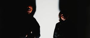 Chris Ryan Williams and Ka Baird look into the camera, faces half-lit in dark clothing, a strip of bright white wall between them.