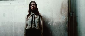 Nina Winder-Lind stands against a stained plaster wall in a pleated white shirt and thin black tie, her shadow beside her, gazing at the camera.